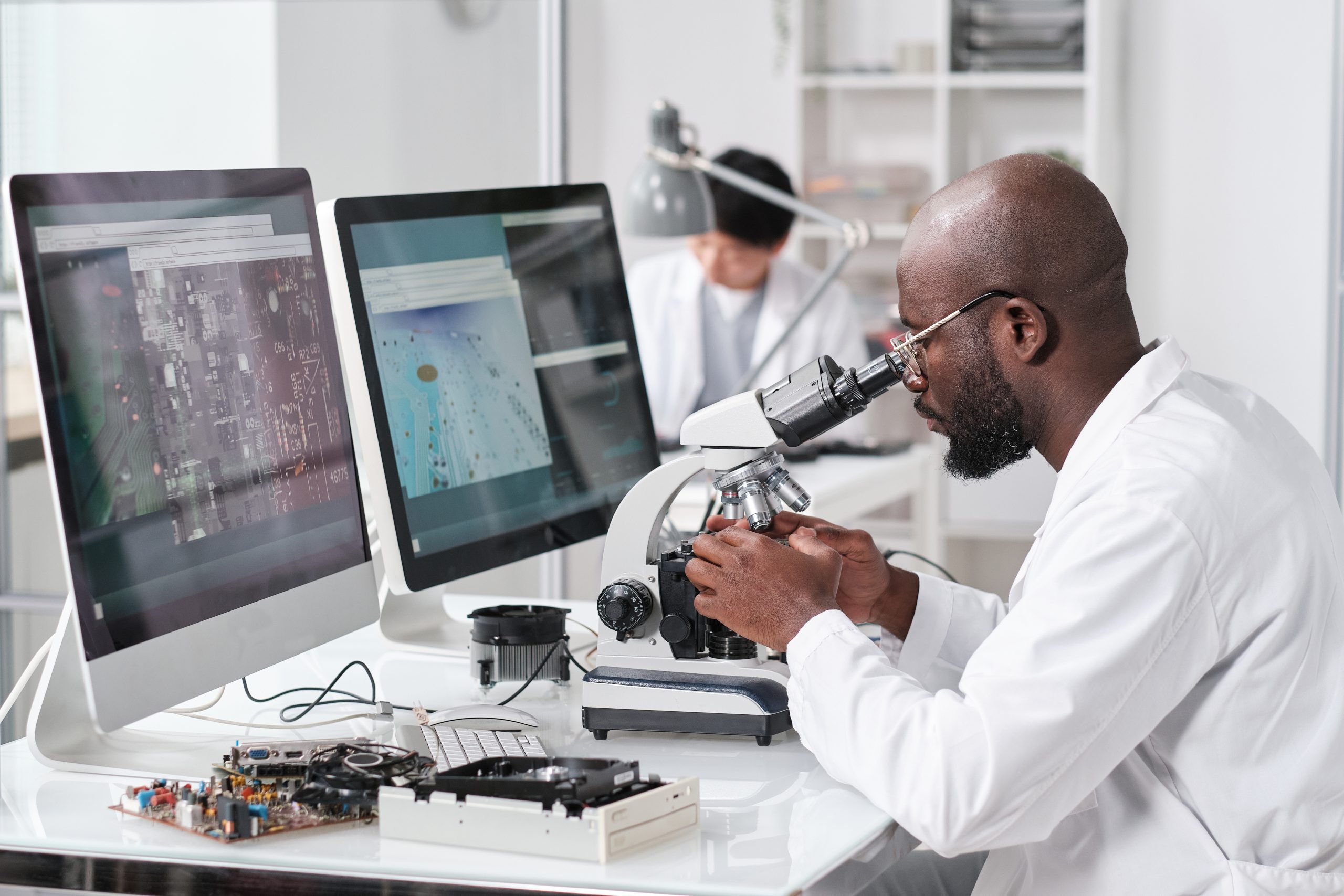 Young serious African man with microscope sitting by workplace in front of computer monitors and colleague on background